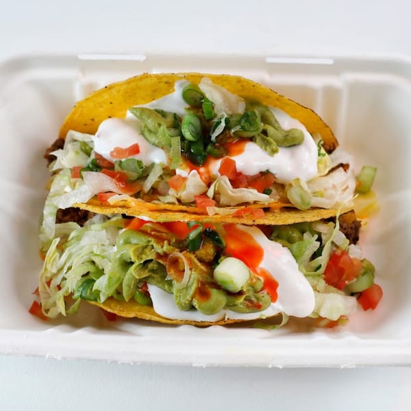 Two Vegan Mob tacos in a white container, topped with lettuce, tomatoes, guacamole, sour cream, and hot sauce.