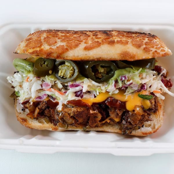 Vegan Mob's vegan BBQ sandwich with coleslaw, jalapeños, and sauce in a white container.