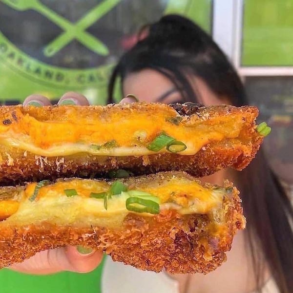 A person holds a crispy, golden-brown vegan grilled cheese sandwich with melted orange cheese and green onions.